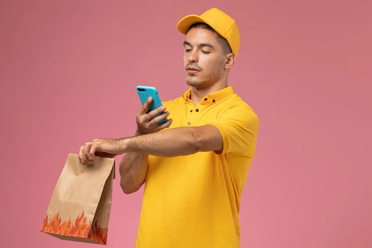 Homem com uniforme amarelo de entregador segurando um saco de papel e verificando informações no celular contra um fundo rosa.