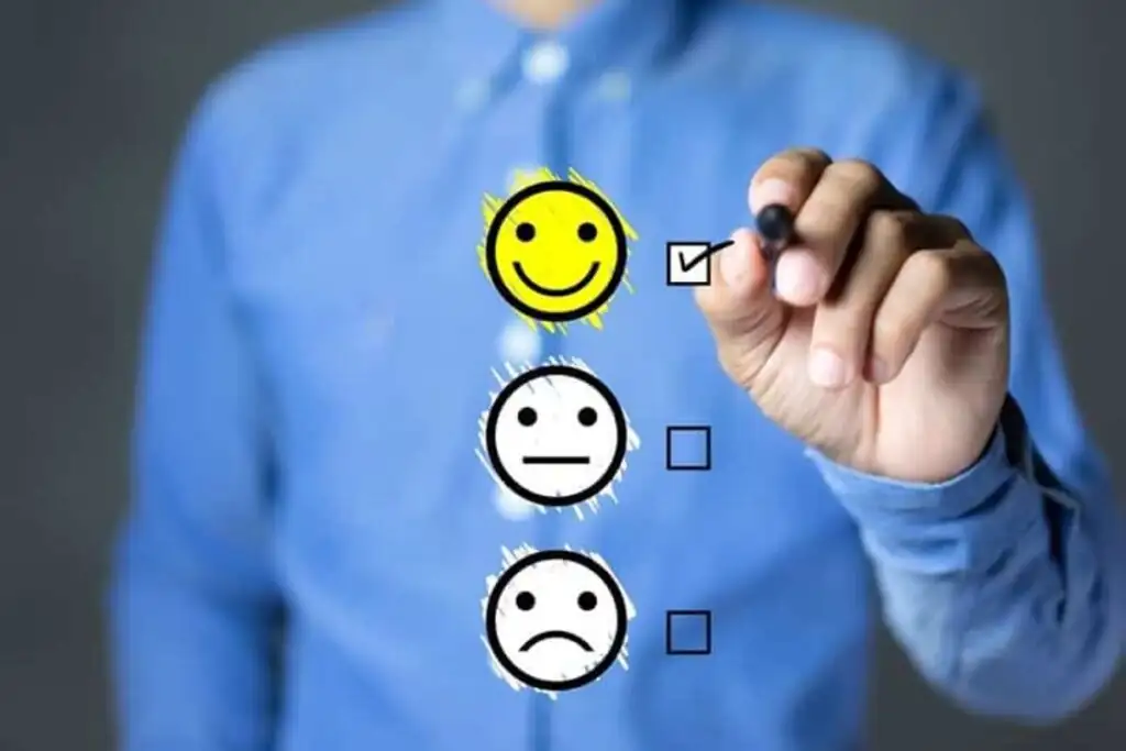 Mão marcando uma caixa de seleção ao lado de um emoji sorridente, em uma escala de satisfação. A cena simboliza o feedback de clientes e a lealdade à marca.