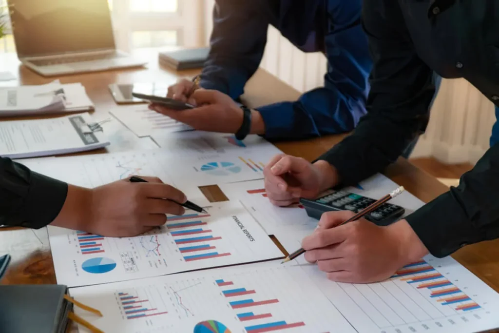 Mãos de profissionais de negócios analisando relatórios financeiros e gráficos de vendas em uma mesa, focando no cálculo de markup.