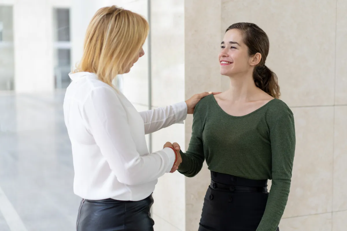 Duas mulheres sorrindo em um aperto de mão e um toque no ombro, em um ambiente interno moderno. A imagem simboliza a construção de um forte "relacionamento com o cliente", confiança e apoio.
