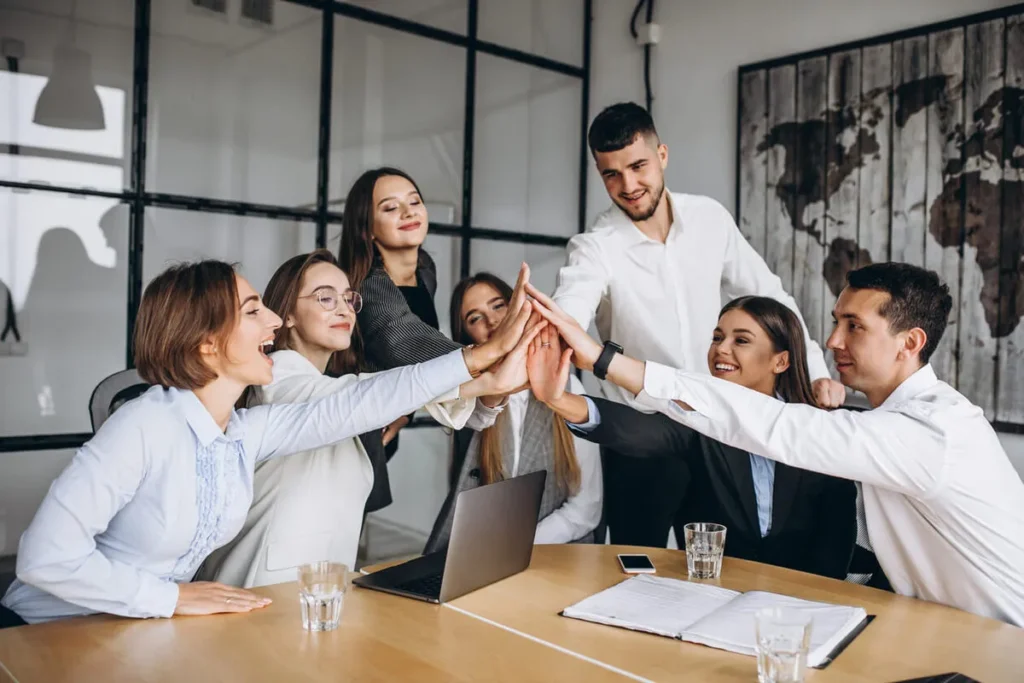 Um grupo de sete profissionais em uma reunião de escritório fazendo um "high five" em equipe.