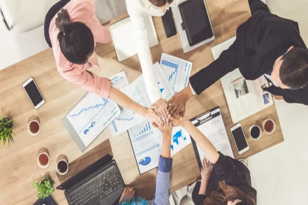 Vista de cima de um grupo de trabalho em um escritório, empilhando as mãos em equipe sobre documentos e um laptop.