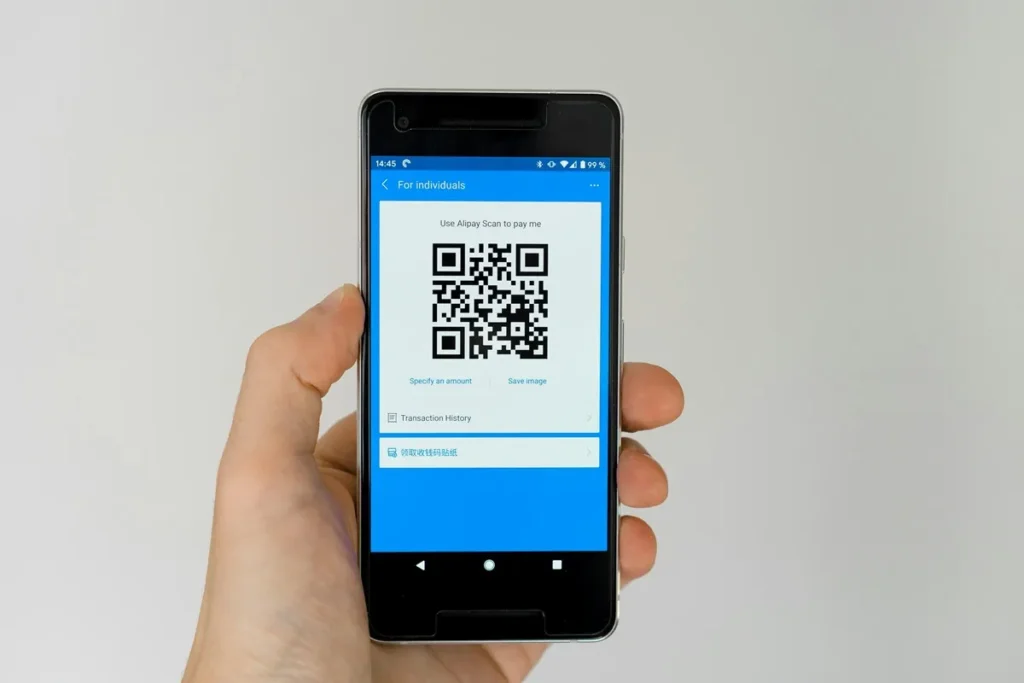 Mão segurando smartphone com tela exibindo QR Code de pagamento no aplicativo Alipay, representando transações digitais seguras.