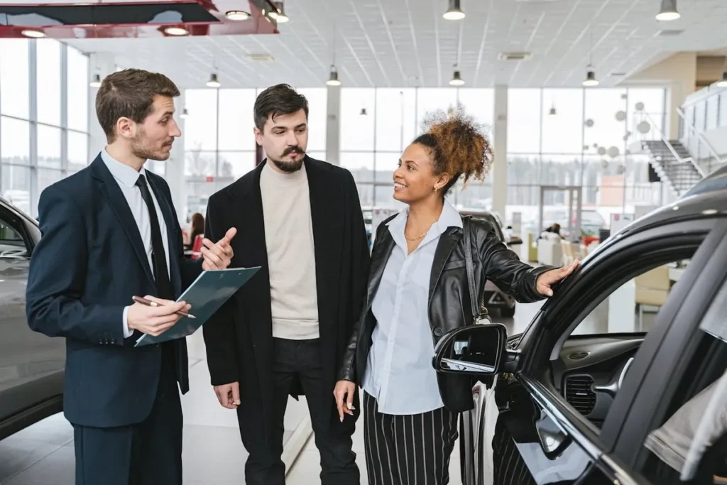 Um consultor de vendas num concessionário de automóveis, de fato e com uma prancheta, a conversar com um casal ao lado de um carro preto. A imagem representa o papel de um consultor de vendas durante a compra de um automóvel.