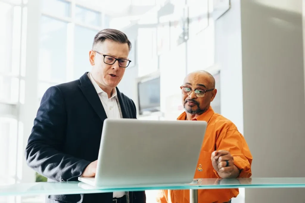 Dois homens, um de fato e outro de camisa laranja, a olhar para um portátil numa mesa de vidro num ambiente de escritório. A imagem simboliza a relação entre um consultor de vendas e um cliente e a sua colaboração.