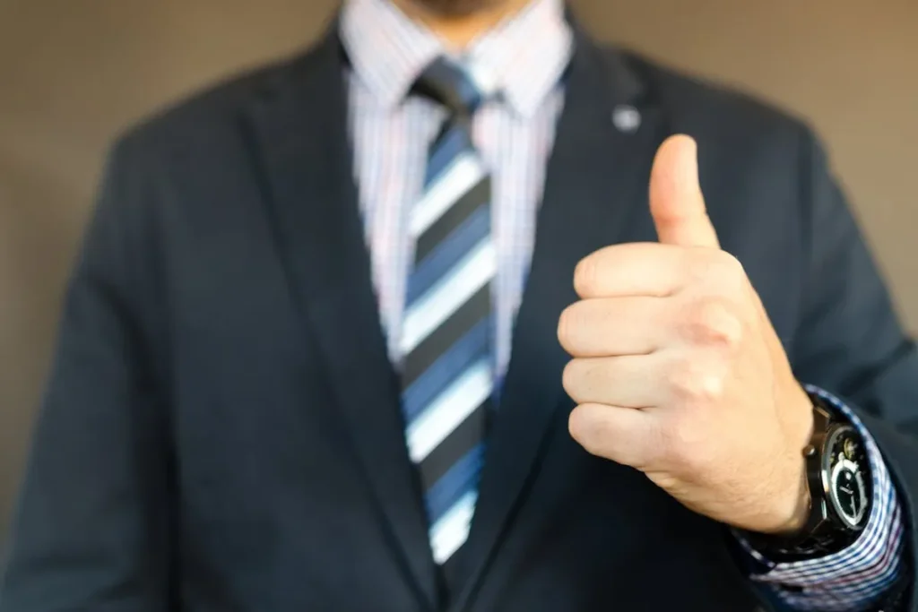 Close-up de um homem de fato, gravata e camisa às riscas, a fazer o gesto de "polegar para cima", a simbolizar sucesso, acordo e confiança. A imagem representa um consultor de vendas bem-sucedido.