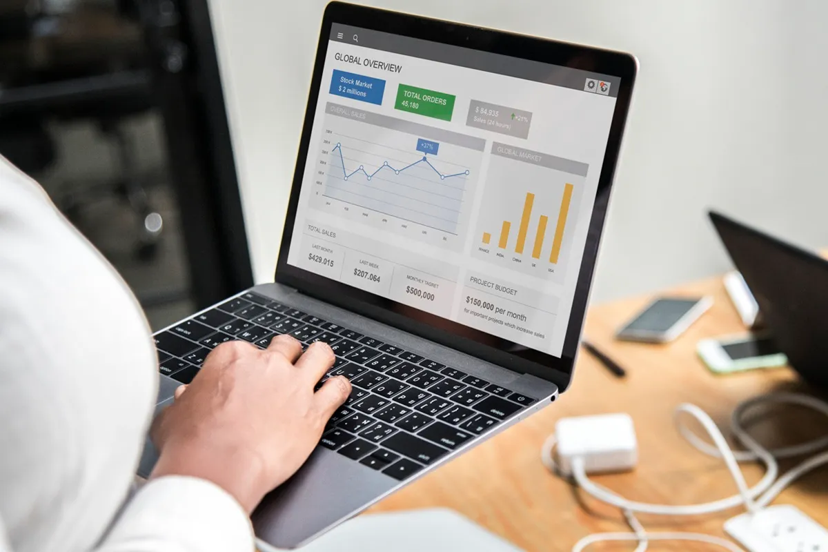 Pessoa usando notebook com dashboard exibindo gráficos de linha e barras, métricas de vendas, pedidos e orçamento de projetos.