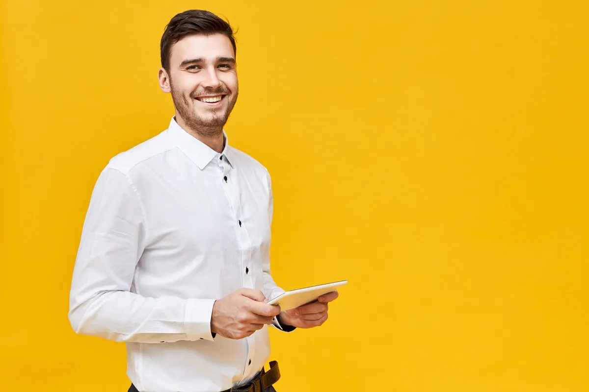 Homem com camisa social branca segurando um tablet e sorrindo, em frente a um fundo amarelo