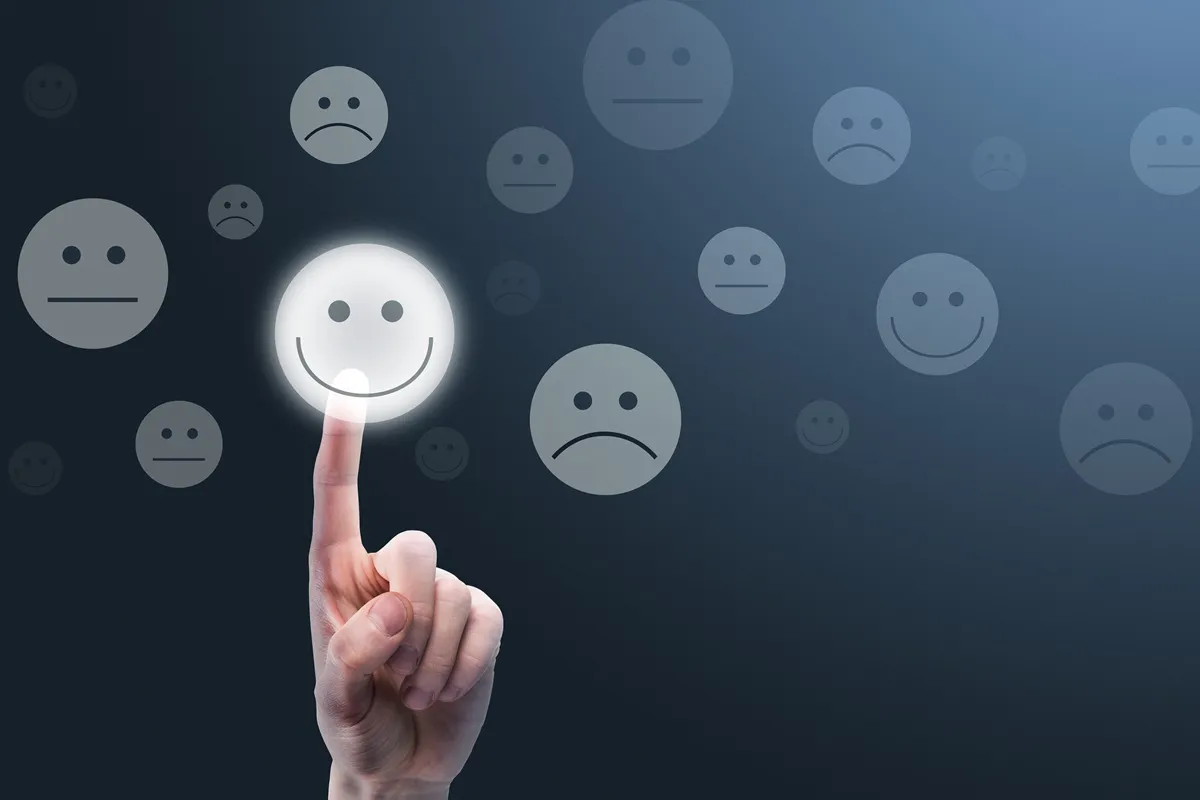 Mão tocando um rosto sorridente em tela escura com ícones de emoções, representando avaliação positiva de atendimento.