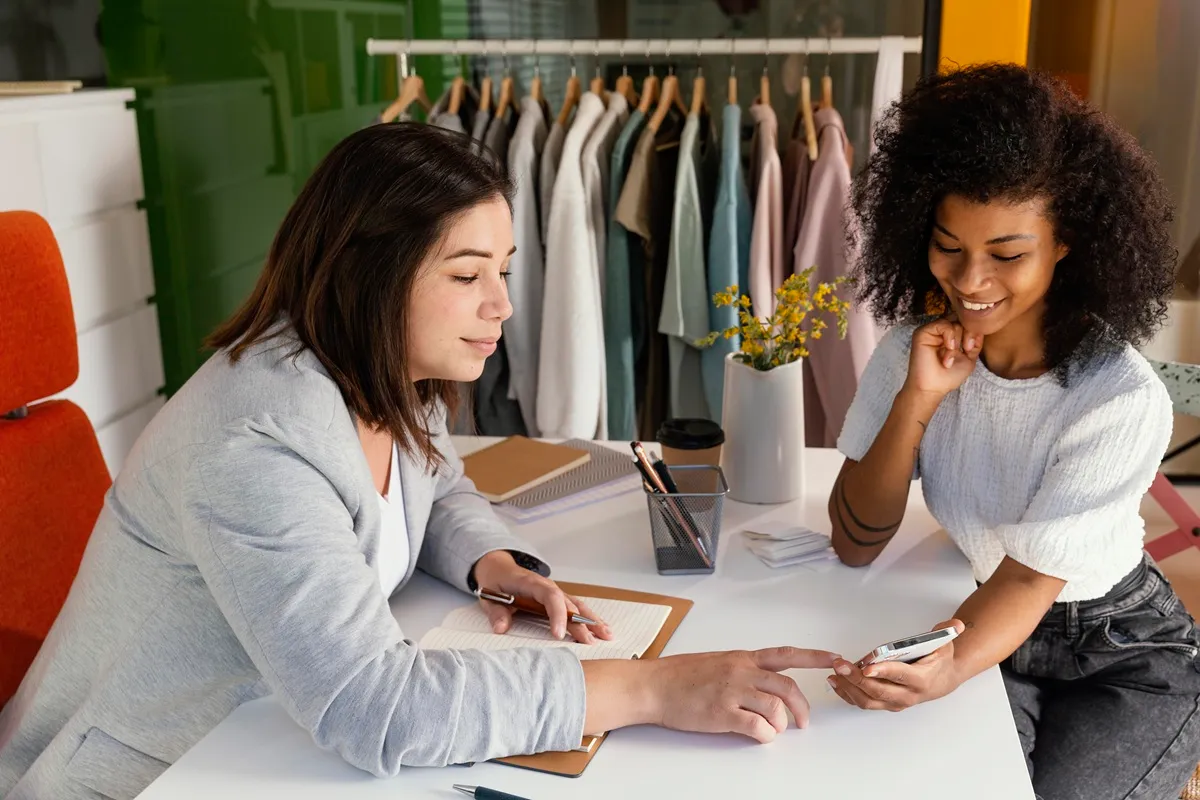 Duas mulheres conversando em um escritório moderno sobre um plano de negócios, com roupas penduradas ao fundo