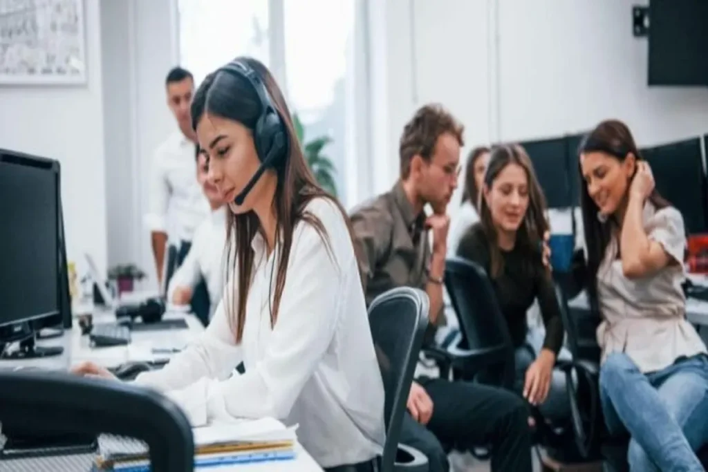 Mulher em um call center usando um headset, operando um computador, enquanto colegas trabalham ao fundo, ilustrando a rotina de pré-venda.