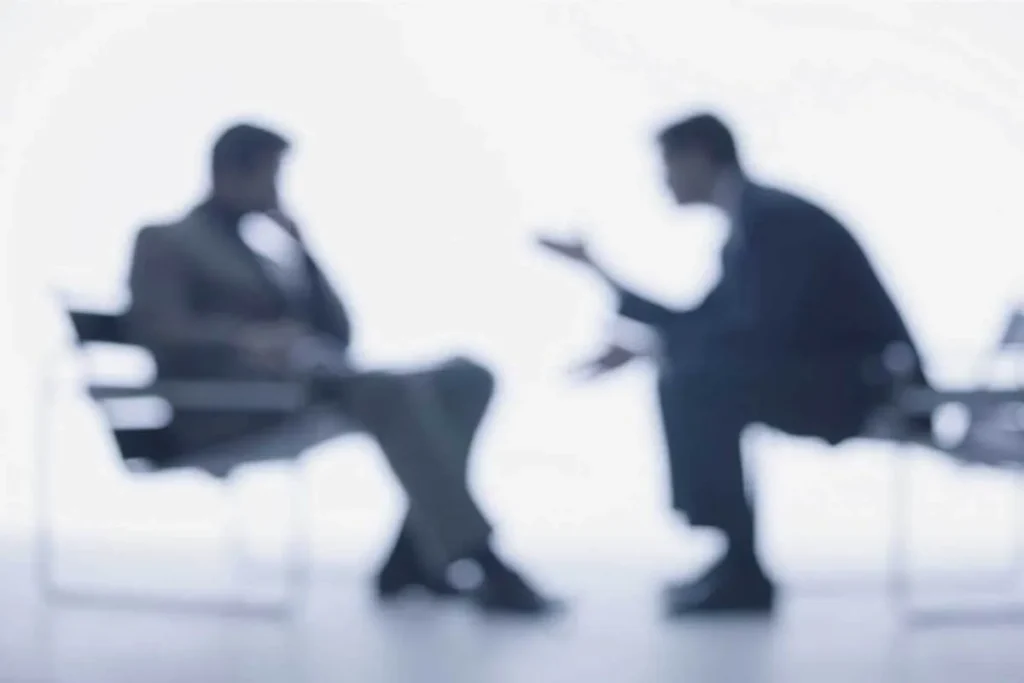 Duas figuras masculinas, desfocadas, sentadas frente a frente, conversando. A imagem sugere uma reunião de pré-venda ou negociação.