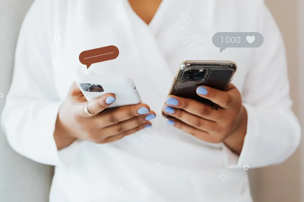 Close-up das mãos de uma pessoa segurando dois smartphones; um com um balão de fala marrom indicando uma mensagem, e o outro mostrando mil curtidas (1000) e um ícone de coração, representando prova social online.
