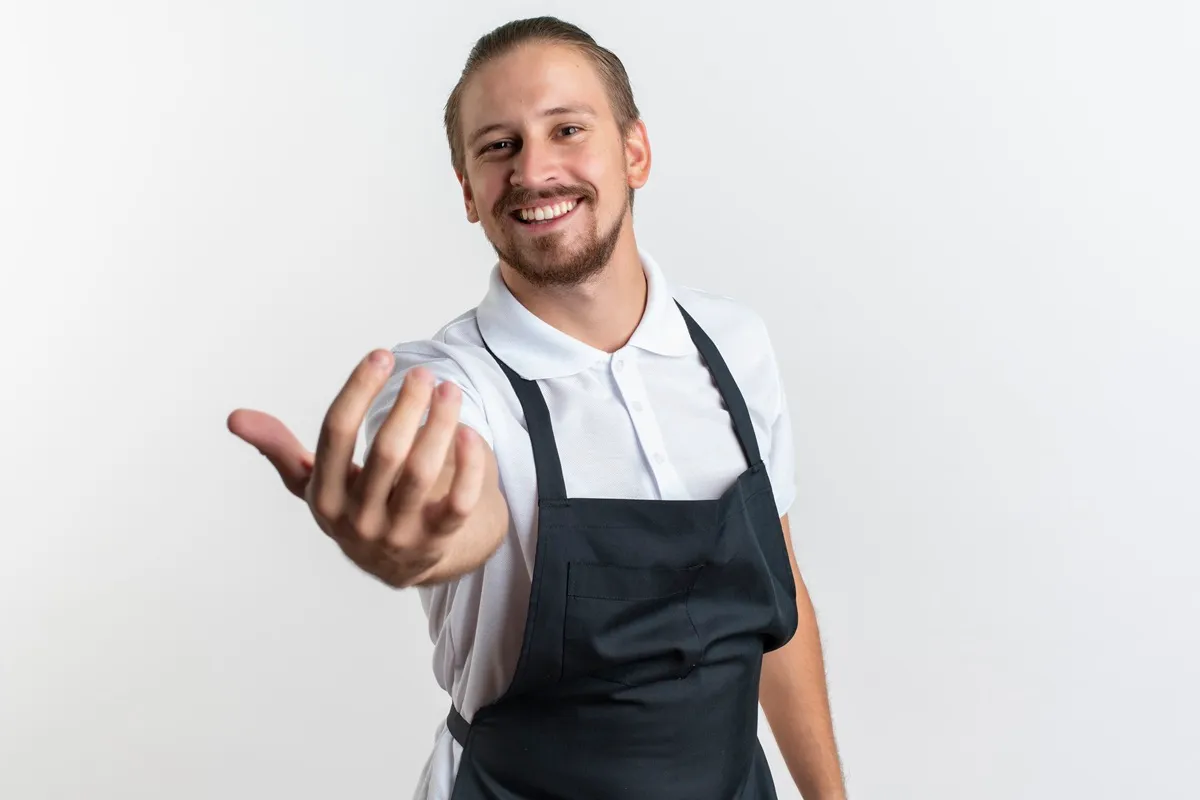 Homem sorridente vestindo uma camisa branca e um avental preto, estendendo a mão para cumprimentar ou oferecer um serviço. Ele está posando para a foto em um fundo branco, transmitindo simpatia e atitude profissional.