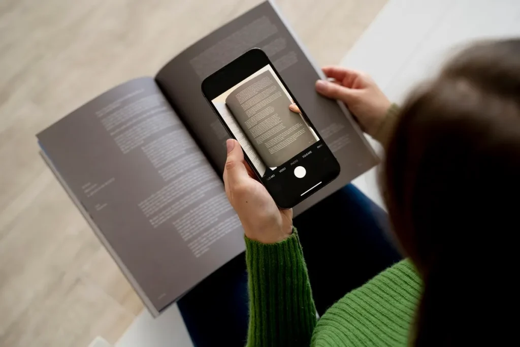 Pessoa usando um smartphone para digitalizar uma página de livro aberta, criando um catálogo digital. Ela está com um suéter verde e a imagem foca no processo de captura de informações do livro.