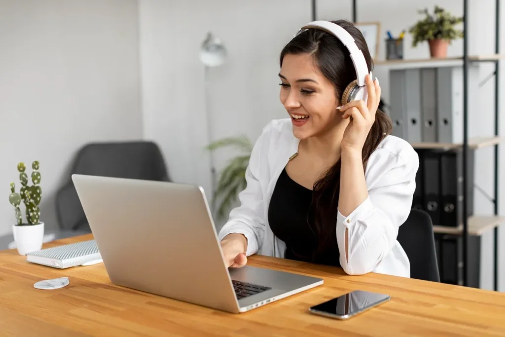 Mulher sorridente usando fones de ouvido enquanto trabalha em um laptop, com um ambiente de escritório moderno