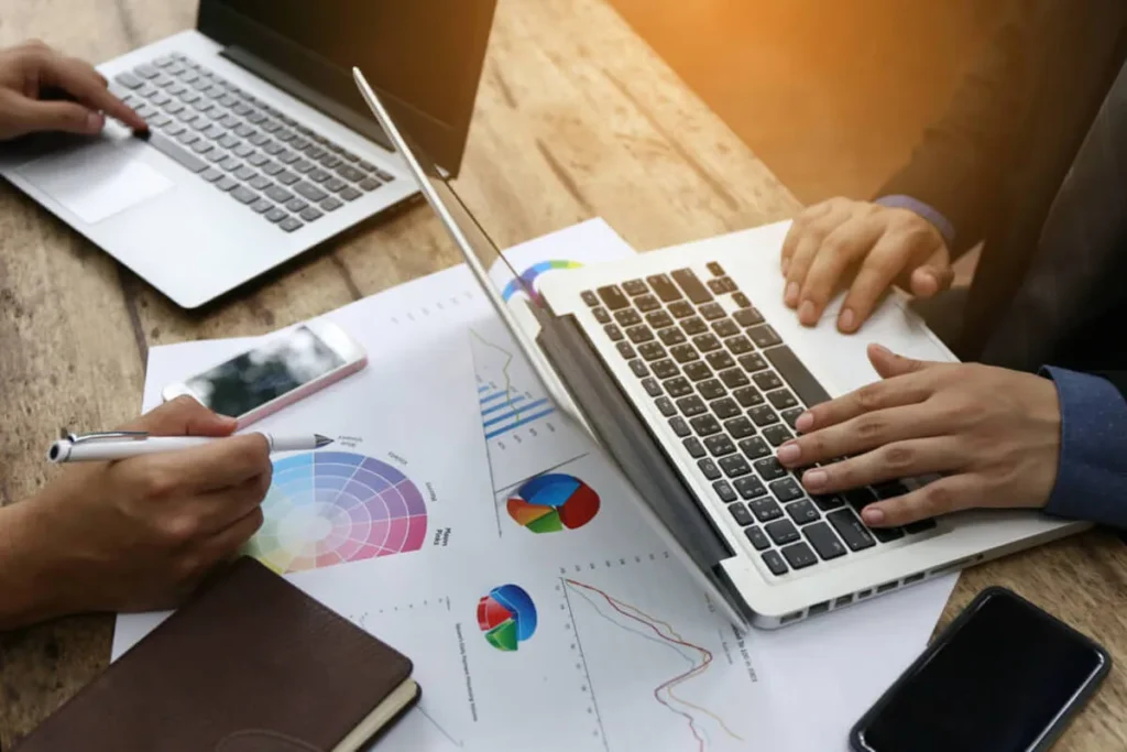 Duas pessoas em uma mesa, usando laptops e analisando documentos e gráficos de benchmarking, com caneta e celulares ao lado.