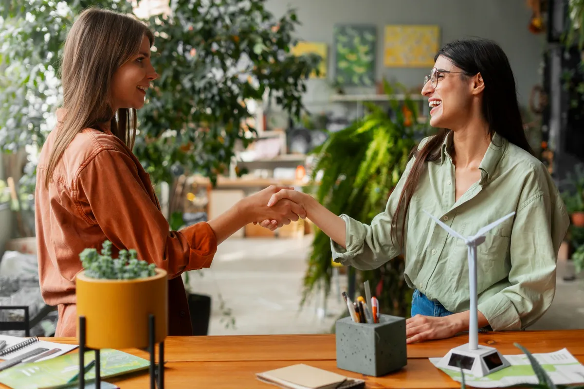 Duas mulheres sorrindo e apertando as mãos em um escritório com temática de sustentabilidade, com plantas e uma maquete de turbina eólica na mesa. A imagem representa "bom atendimento ao cliente" e colaboração.