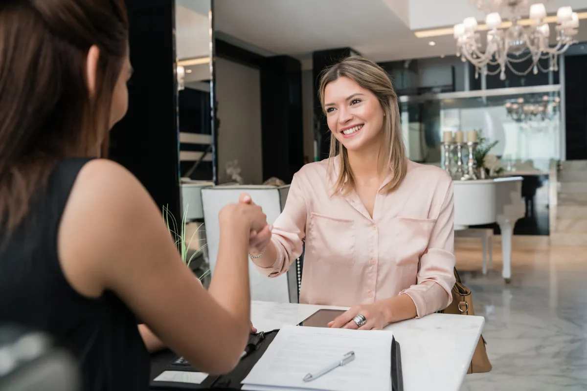 Duas mulheres se cumprimentando com um aperto de mão e sorrindo, em um ambiente elegante que parece ser a recepção de um hotel ou escritório. A imagem transmite a ideia de "bom atendimento ao cliente", parceria e satisfação.