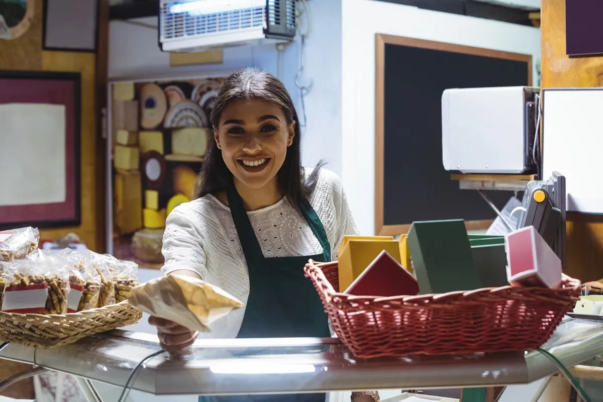 Mulher sorrindo e entregando um pacote a um cliente em uma loja de varejo, com cestas de produtos e itens coloridos ao fundo.