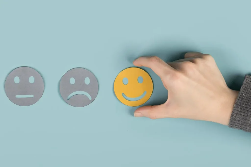Mão selecionando um ícone de rosto sorridente, representando a mudança de um estado emocional, com rostos neutro e triste ao lado