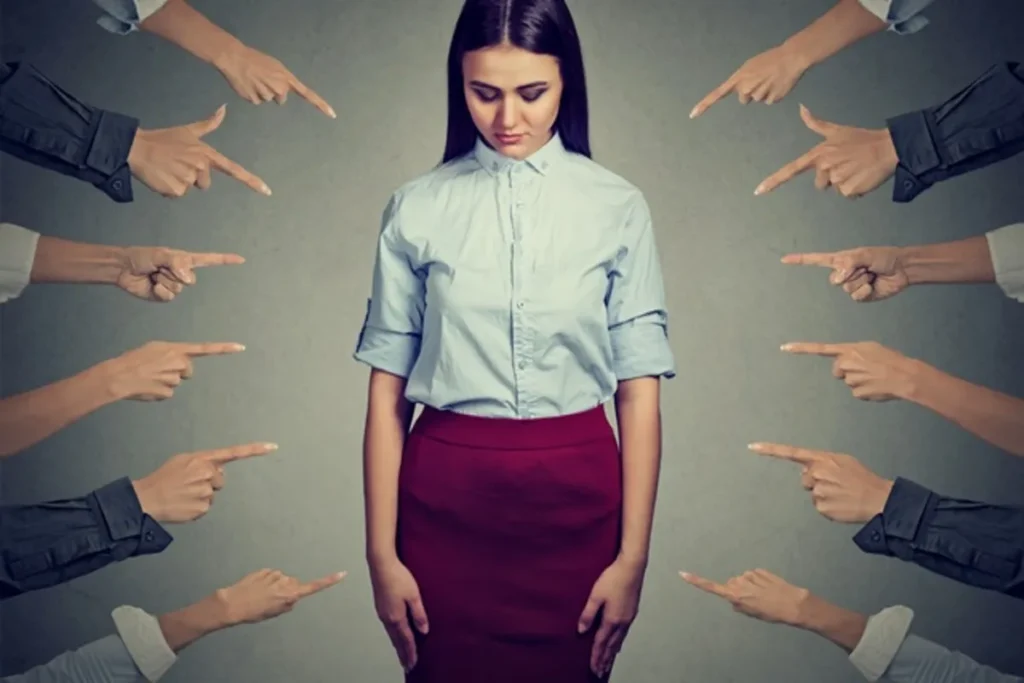 Mulher em blusa azul e saia bordô sendo apontada por várias mãos em círculo, representando a pressão do julgamento, o Efeito Horn ou o assédio moral e culpa.