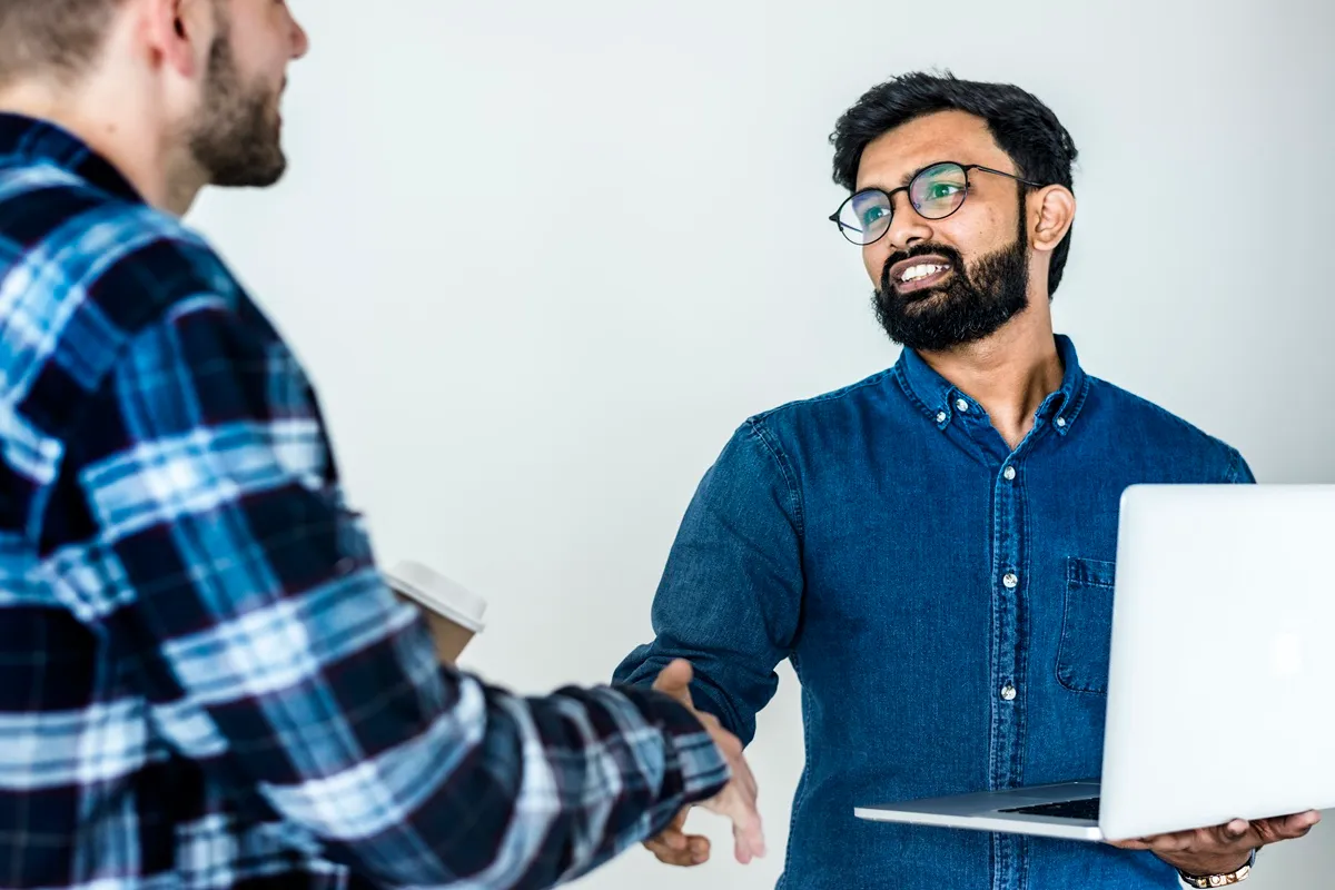 Homem de camisa xadrez apertando a mão de consultor de óculos e camisa jeans que segura um notebook