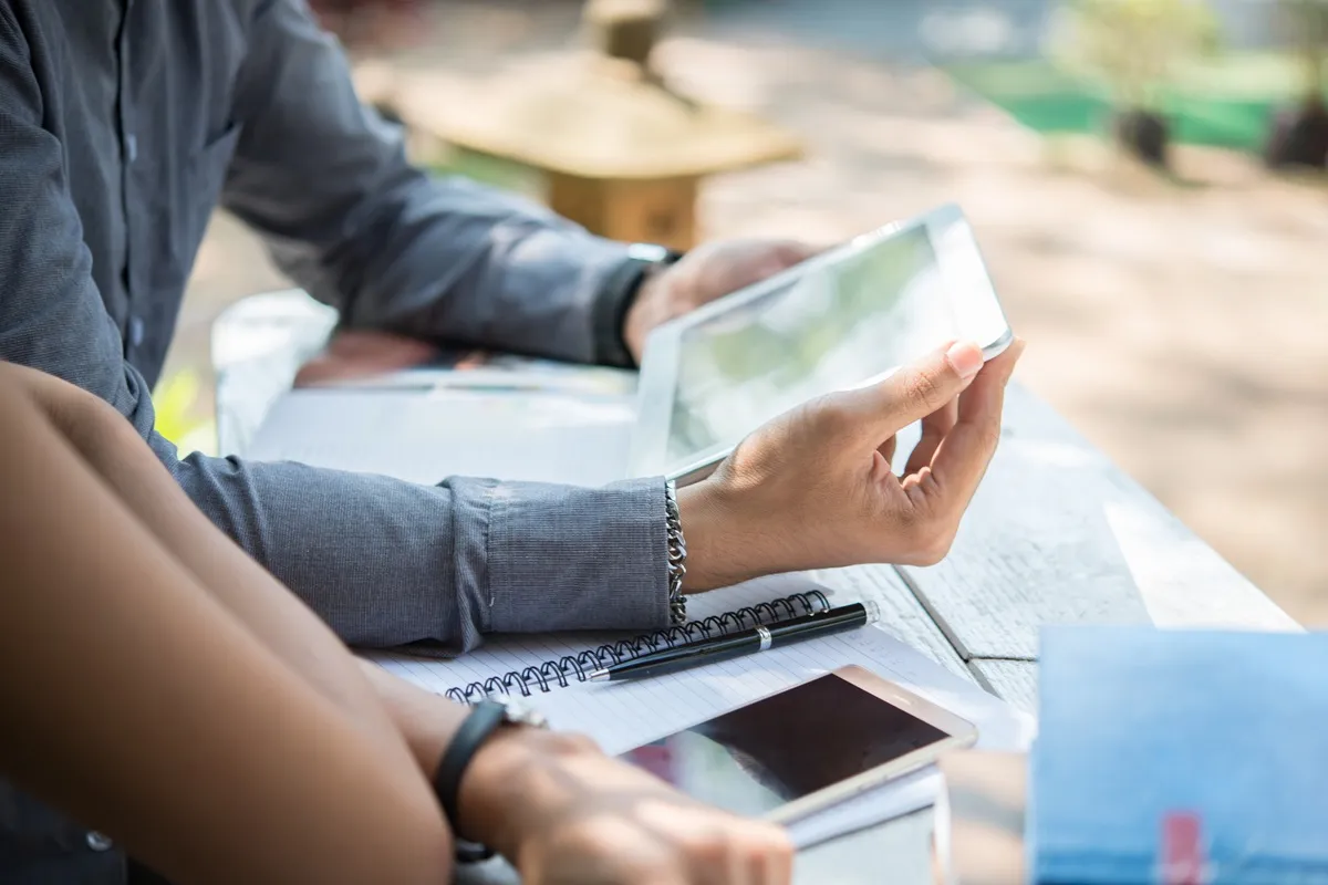 Imagem de duas pessoas usando dispositivos eletrônicos, como tablet e celular, em um ambiente tranquilo e ao ar livre, indicando o conceito de ganhar dinheiro online respondendo pesquisas.