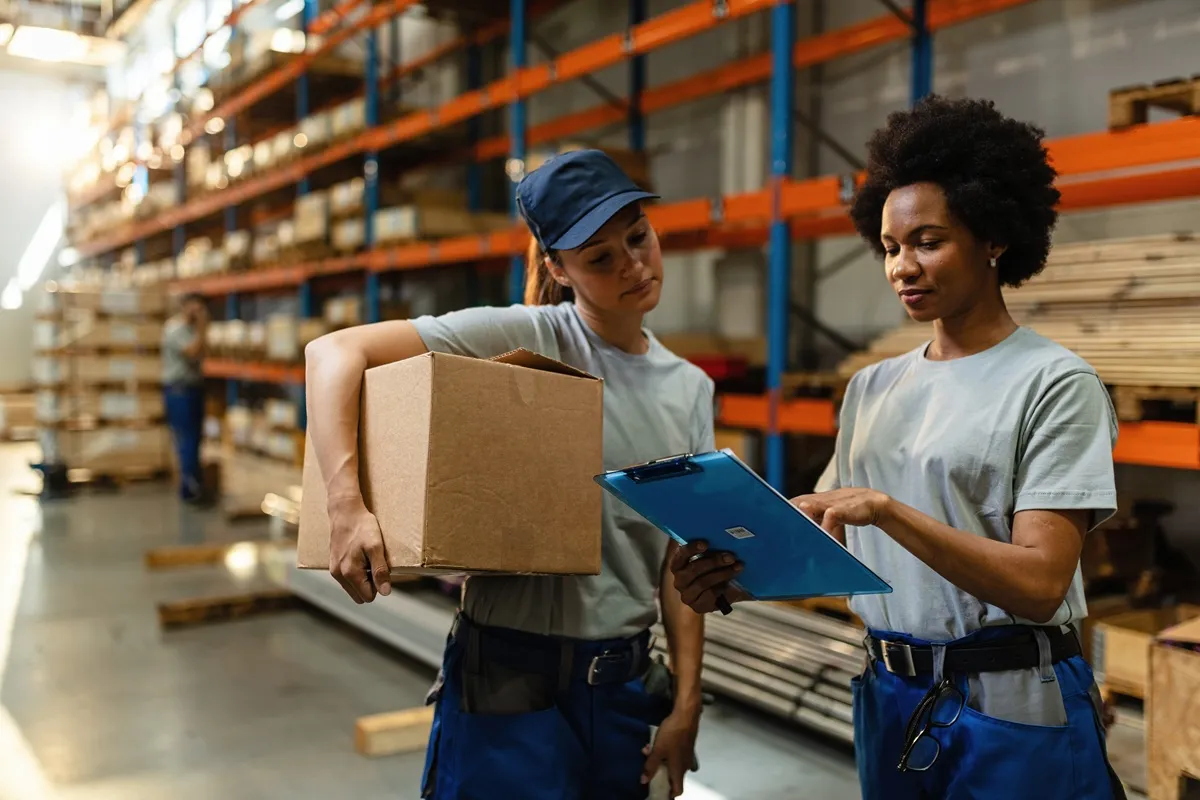 Duas mulheres em uniforme de trabalho conferindo dados em uma prancheta enquanto seguram uma caixa de papelão em um galpão de estoque com prateleiras altas ao fundo.