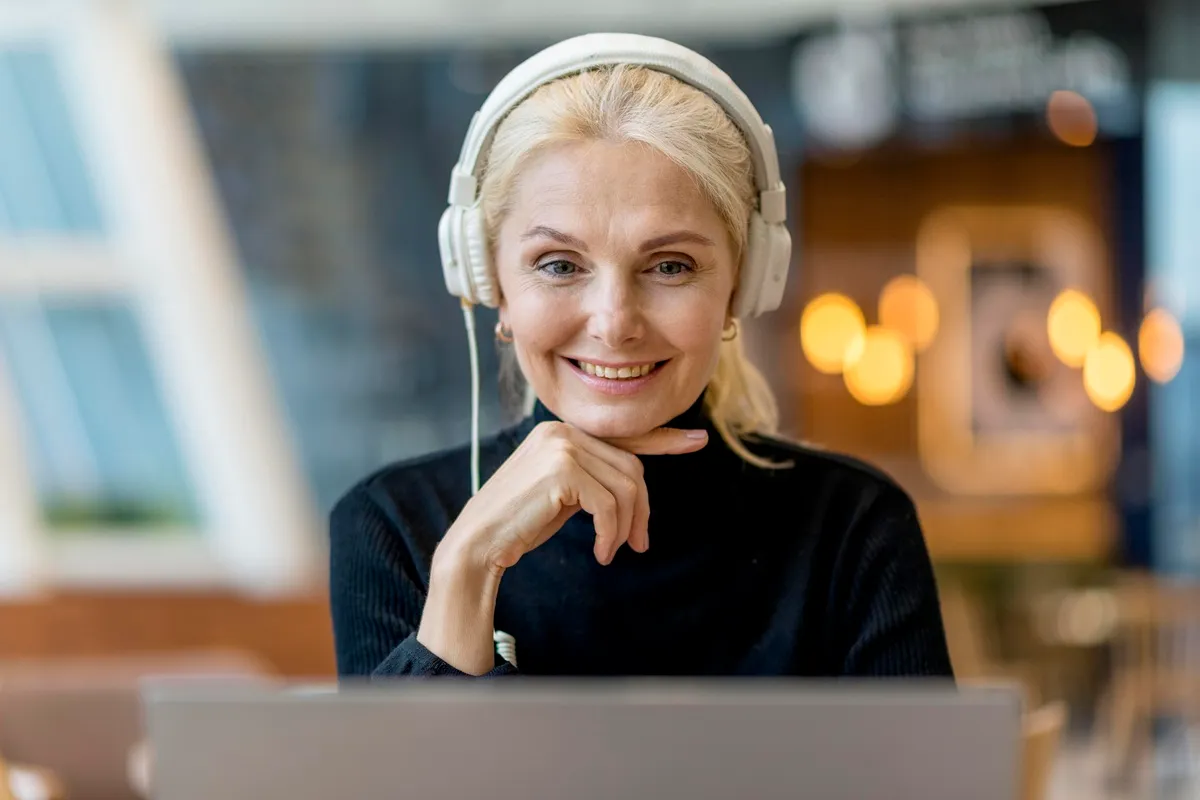 Mulher sorridente com fones de ouvido, sentada diante de um laptop, em ambiente moderno e iluminado