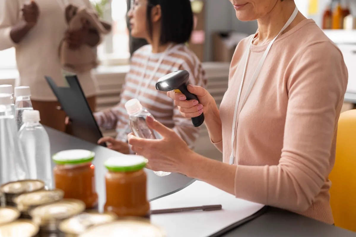 Mulher escaneando garrafa de plástico com leitor de código de barras em ponto comercial, enquanto outra pessoa trabalha ao fundo no computador. O ambiente é organizado com produtos expostos sobre a mesa.