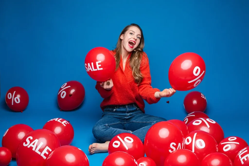 Mulher sorridente segurando balões vermelhos com as palavras SALE e % ao redor, em um cenário de liquidação e ofertas especiais