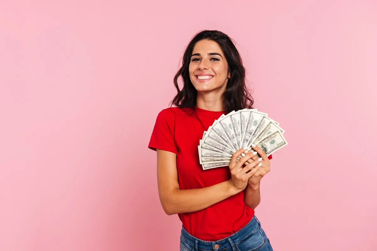 Mulher sorridente de camiseta vermelha segurando leque de notas de dólar contra fundo rosa