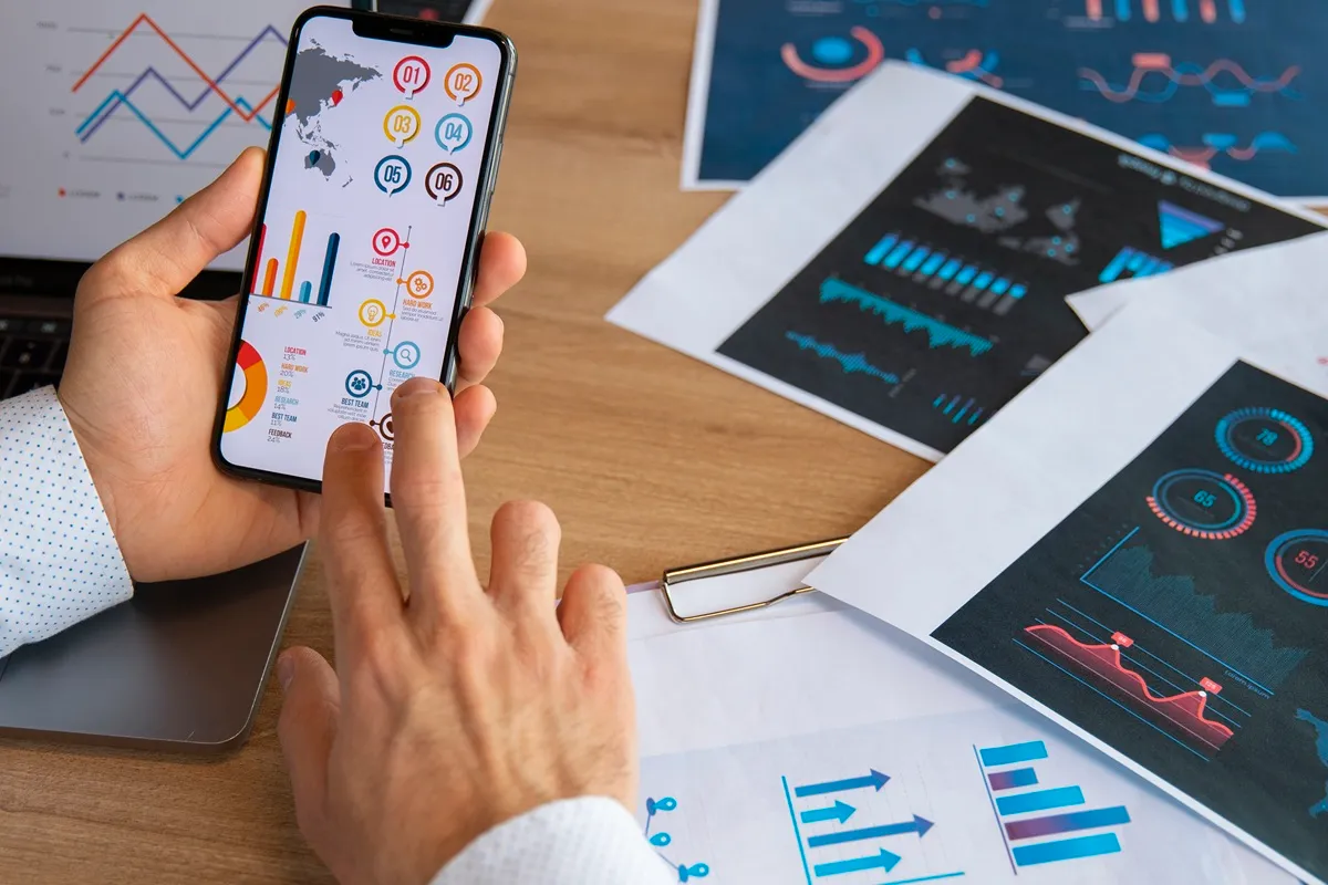 Pessoa usando smartphone com gráficos coloridos e indicadores de desempenho, rodeada por folhas impressas com dashboards e análises visuais.