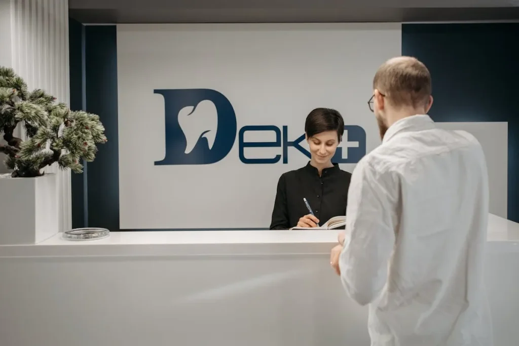 Recepcionista sorridente em balcão de clínica com logotipo de dente, atendendo cliente e focando na experiência do cliente.