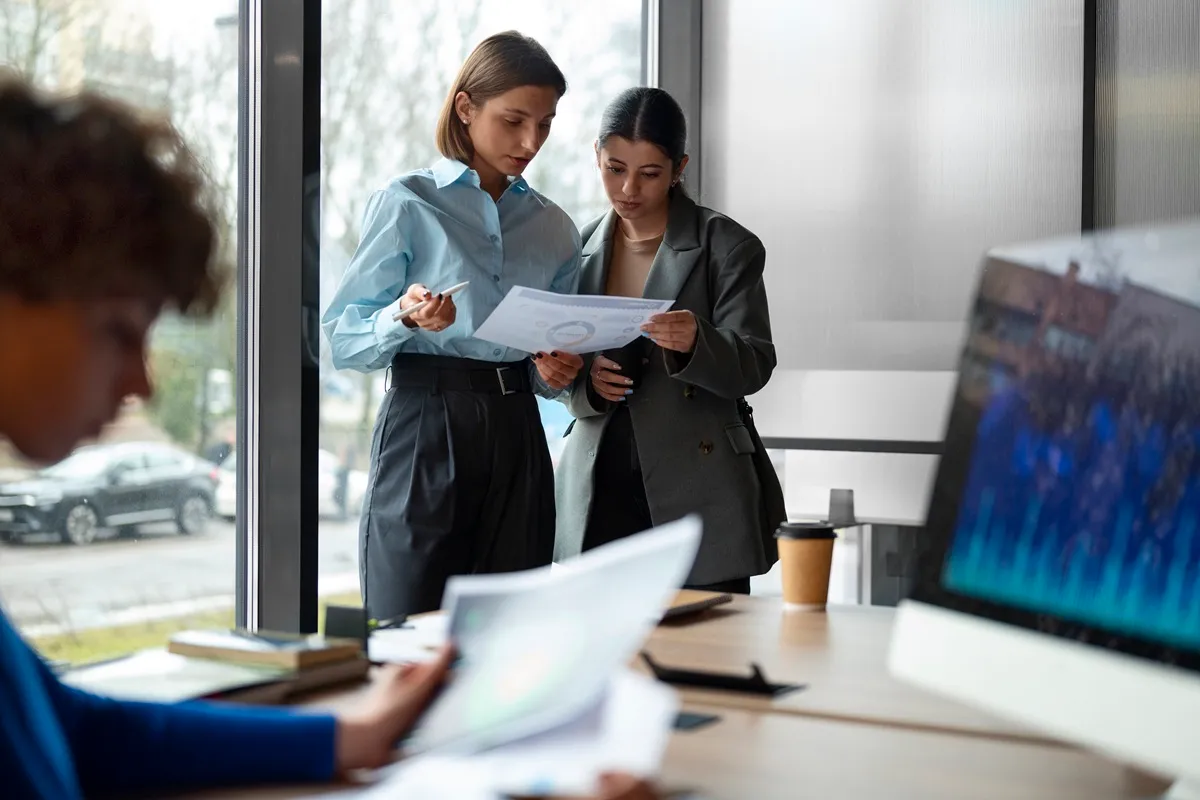 Duas mulheres revisam juntas um relatório impresso de auditoria de processos em frente a uma janela, enquanto um colega ao fundo trabalha em computador