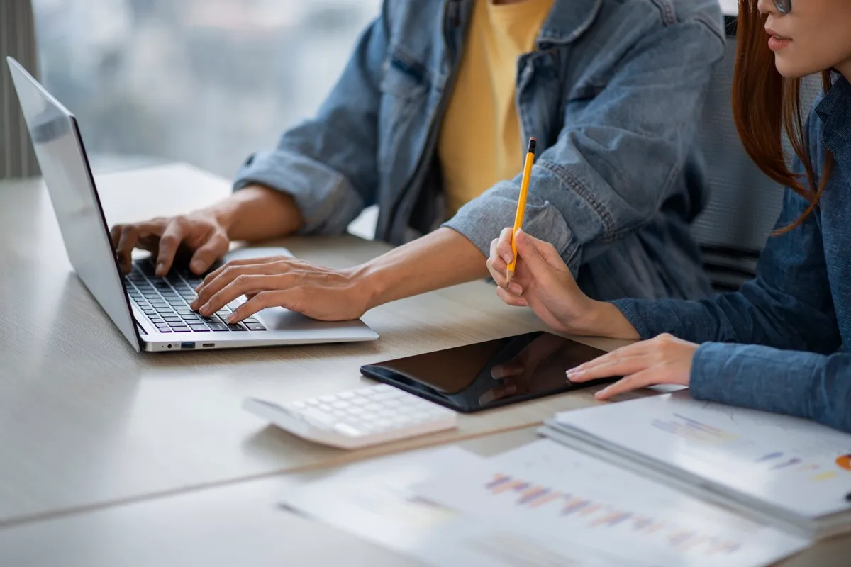 Duas pessoas colaborando em auditoria de processos: uma digita no notebook enquanto a outra segura um lápis sobre um tablet, com relatórios impressos de gráficos à frente