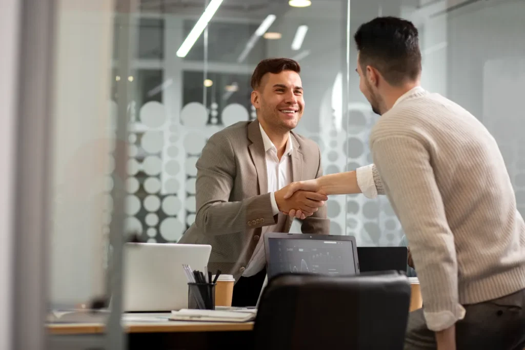 Dois parceiros de negócios apertam as mãos com um sorriso, representando a consistência em parcerias e o fechamento de um negócio.