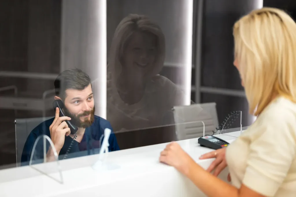 Um recepcionista, representando consistência no atendimento, fala ao telefone enquanto uma cliente aguarda em um balcão de recepção.