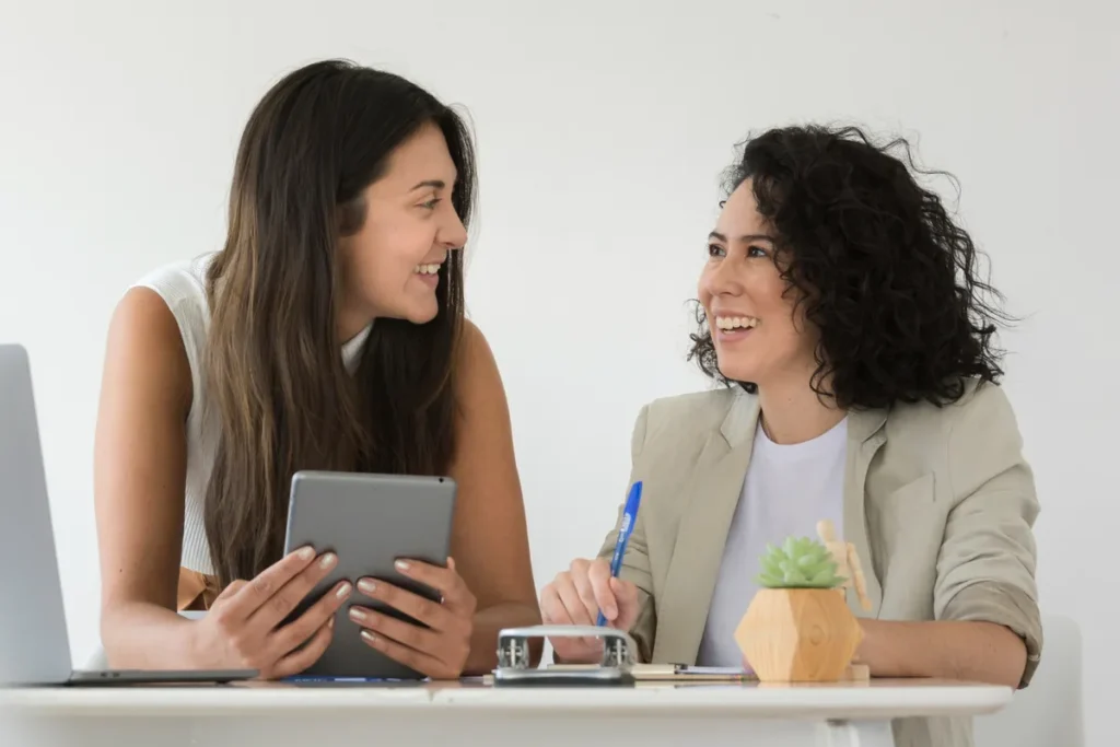 Duas mulheres sorridentes, representando consistência e colaboração, trabalham juntas em uma reunião em um escritório.