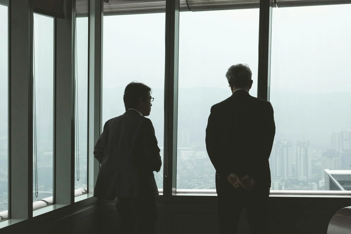 Dois executivos de terno observando a cidade pela janela de um prédio comercial, simbolizando planejamento estratégico ou reunião de negócios.