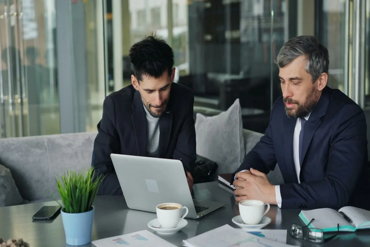 Dois empresários analisando gráficos e informações em um notebook durante uma reunião em ambiente corporativo.