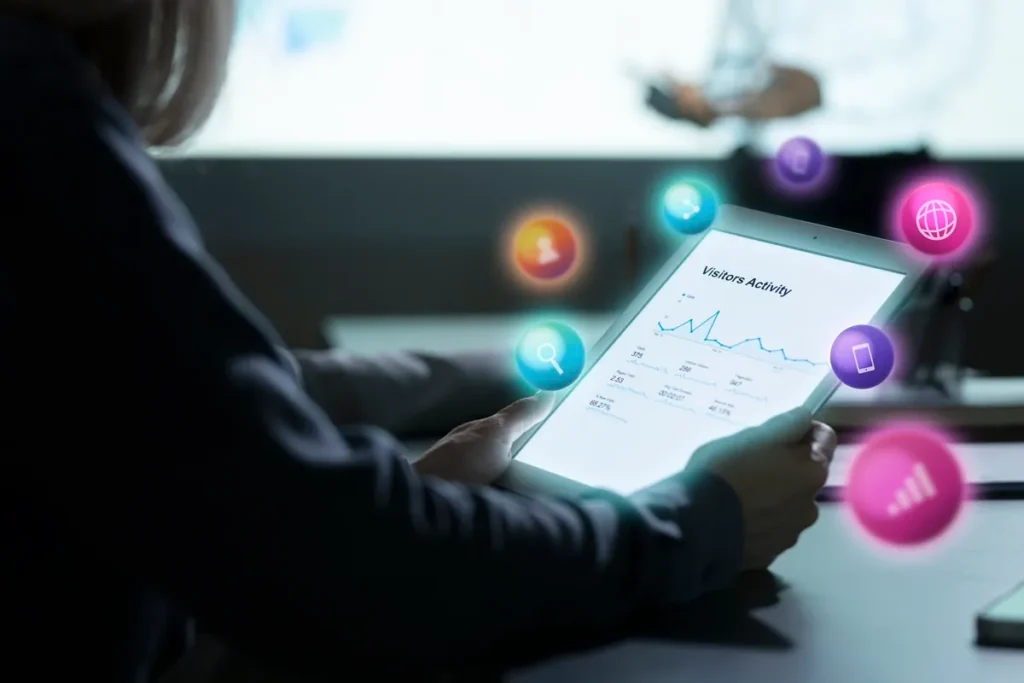 Pessoa analisando gráfico de atividade de visitantes em tablet, cercada por ícones digitais coloridos representando comunicação, conexão e dispositivos móveis.