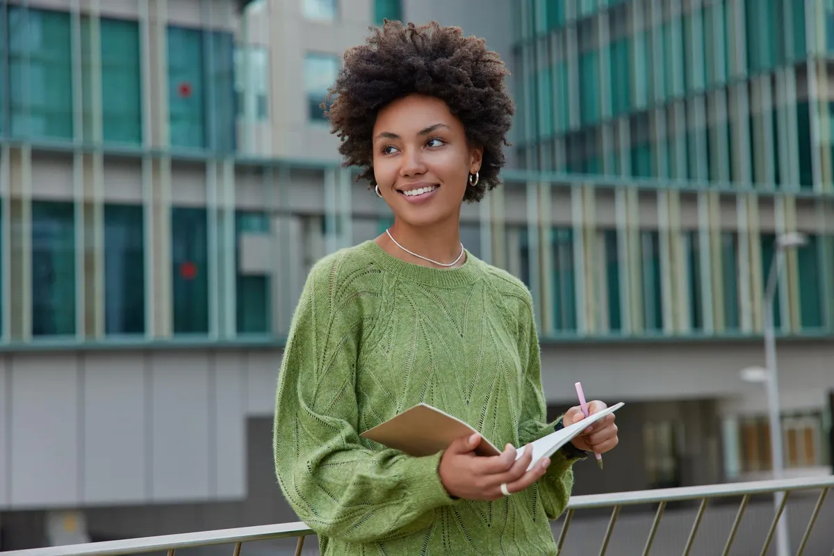 Jovem estudante sorridente ao ar livre com caderno e caneta, em frente a prédio moderno.