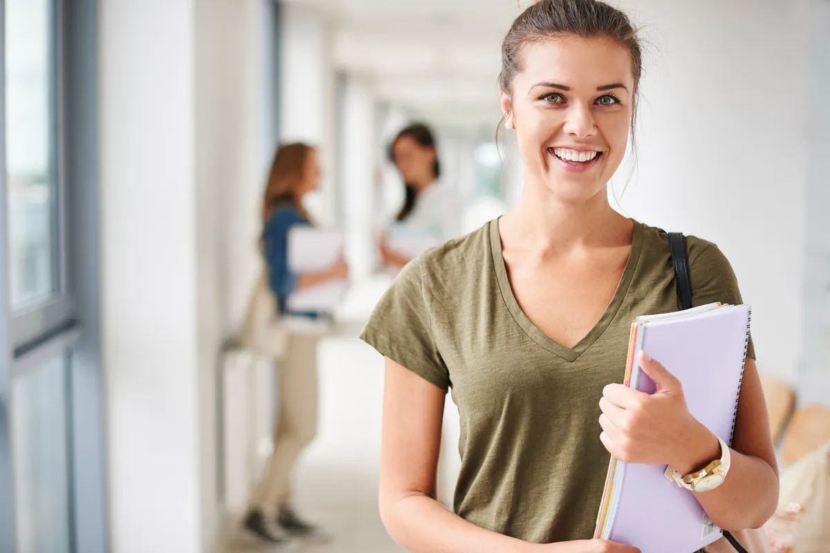 Estudante sorridente segurando cadernos em corredor de escola, com outras alunas conversando ao fundo.