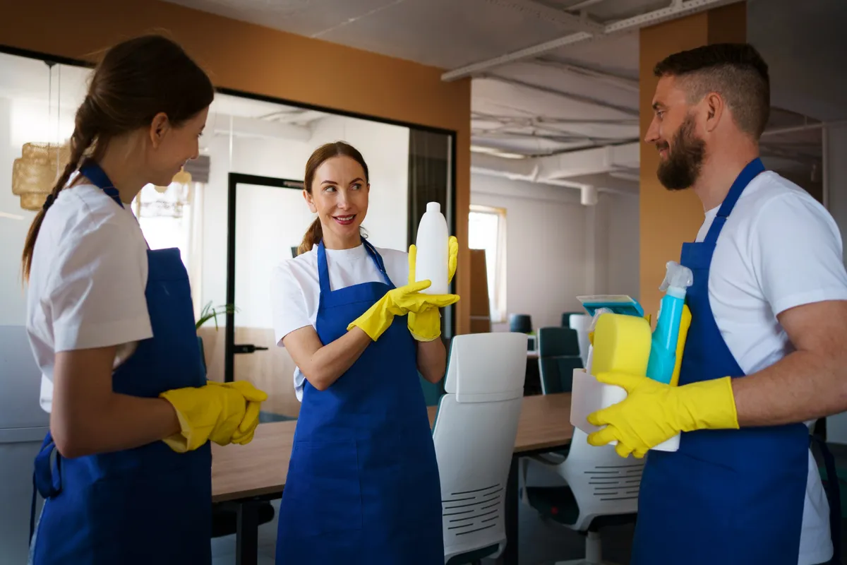 Equipe de limpeza com uniformes azuis e luvas amarelas conversando e segurando produtos de limpeza em ambiente corporativo.