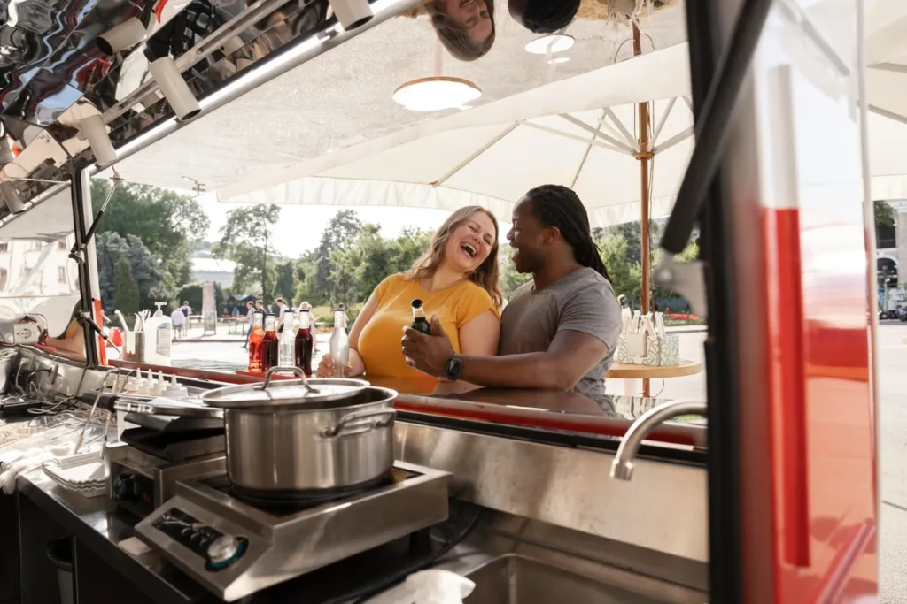 Casal sorrindo e bebendo em um balcão de food truck, com a cozinha visível em primeiro plano.