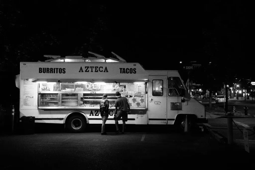 Um food truck branco, com as palavras "Burritos", "Azteca" e "Tacos" escritas na lateral, atendendo a duas pessoas à noite em uma rua escura.