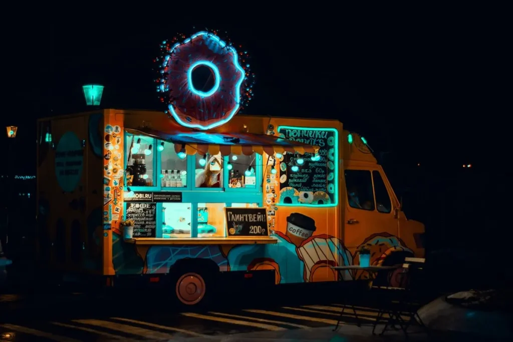 Food truck amarelo e azul com um letreiro de neon em formato de donut, servindo donuts e café à noite.