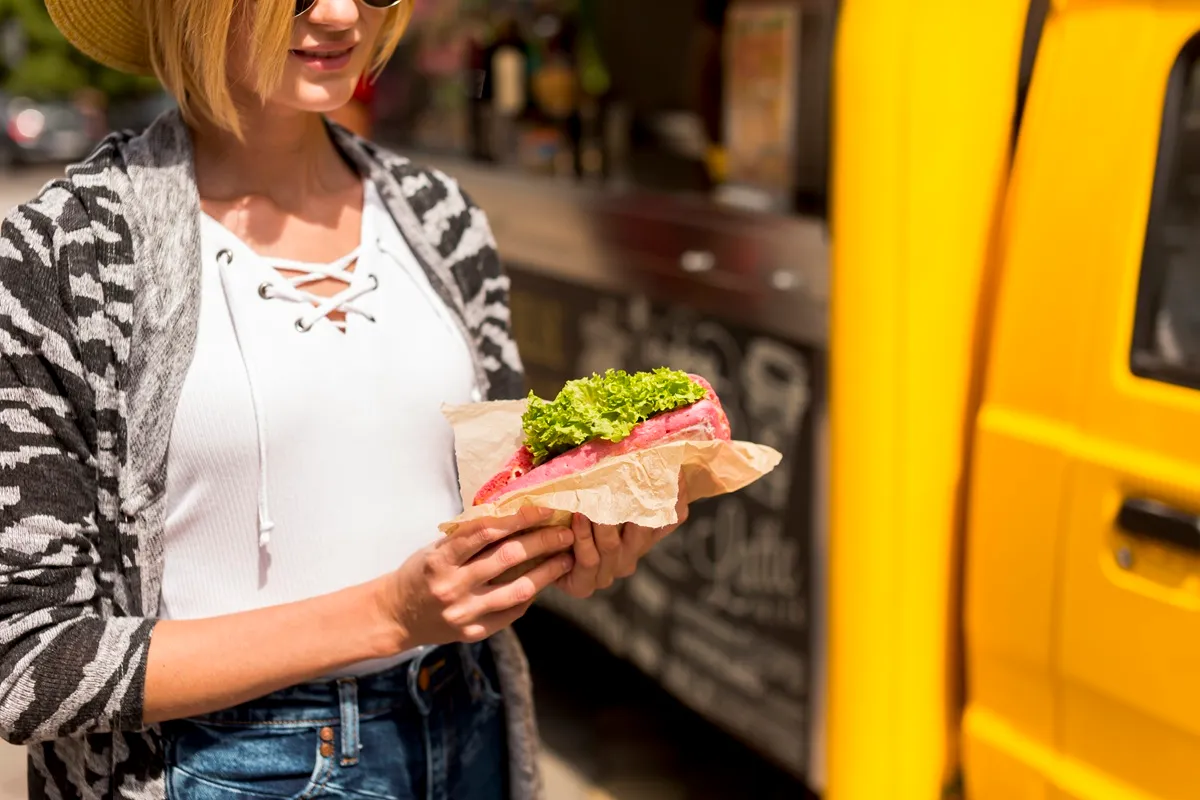 Imagem de uma mulher segurando um lanche com alface em frente a um food truck amarelo. Ela usa óculos escuros e um chapéu, sorrindo enquanto aprecia a comida.
